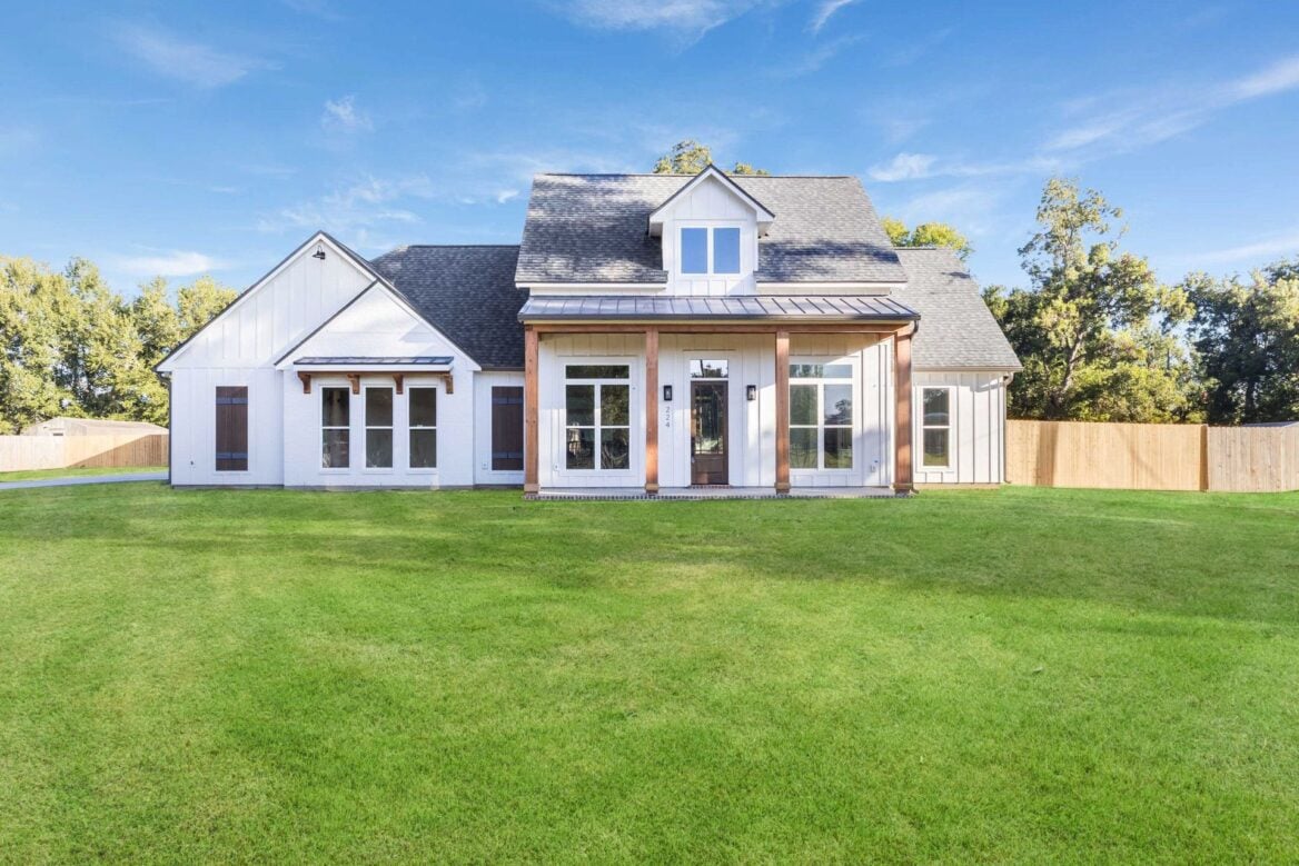 A modern white house with a dark roof, large windows, and wooden posts on the front porch, situated on a spacious green lawn with a wooden fence and trees in the background under a clear blue sky.