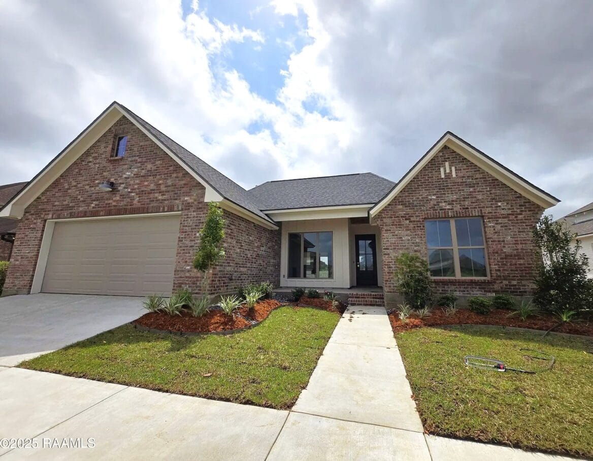 A single-story brick house with an attached two-car garage, large windows, and a covered front entry. The yard has young landscaping, and a concrete driveway and walkway lead to the front door under a partly cloudy sky.