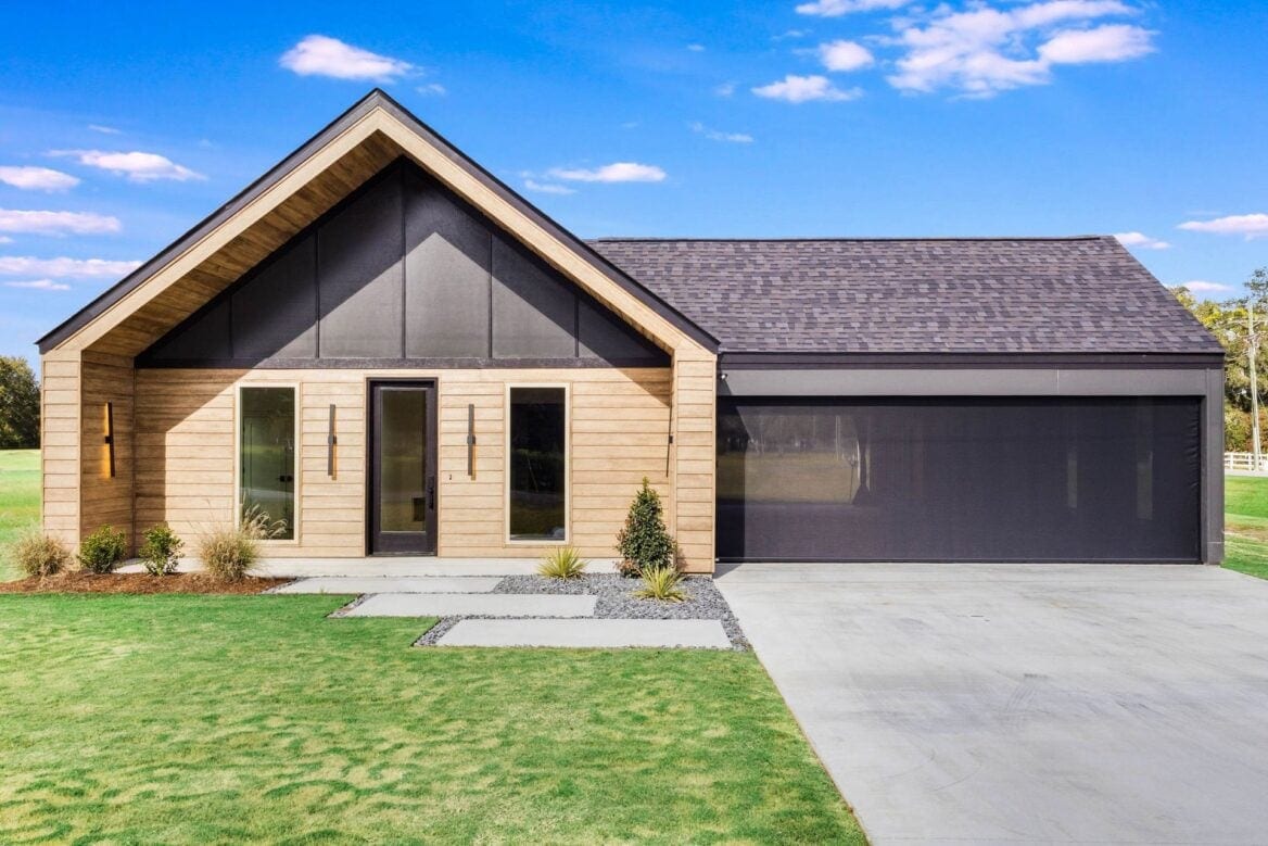 A modern single-story house with wood siding, large windows, a dark gable roof, and an attached two-car garage, set on a lawn with a concrete driveway under a blue sky with scattered clouds.