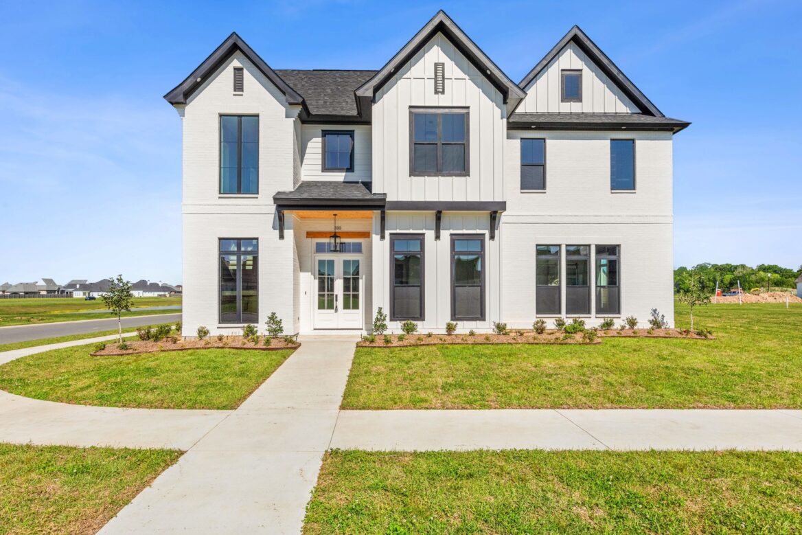 A two-story modern farmhouse-style house with white siding, black trim, multiple large windows, and a front porch, set on a green lawn with a sidewalk leading to the entrance.