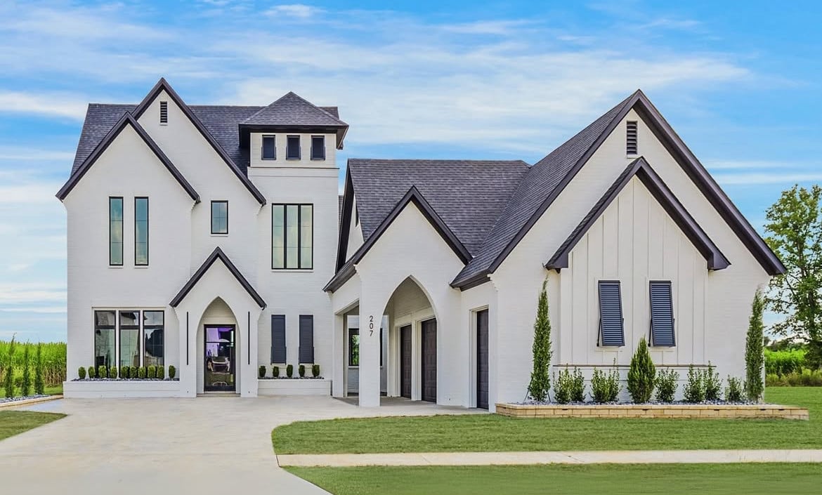 A modern two-story house with white brick exterior, dark roof shingles, and large windows. There is a driveway, green lawn, and minimal landscaping with small shrubs in front of the home.
