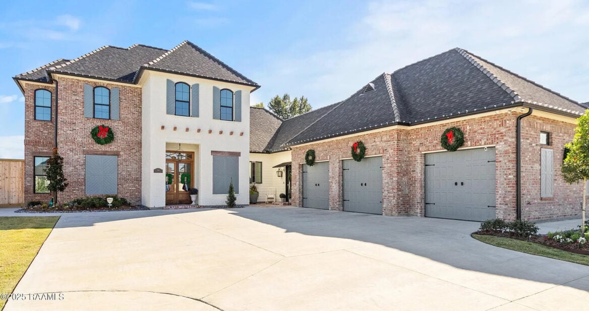 A two-story brick house with an attached three-car garage. Both the house and garage have black roofs and are decorated with holiday wreaths on the doors. The driveway is wide and there is a small landscaped area.