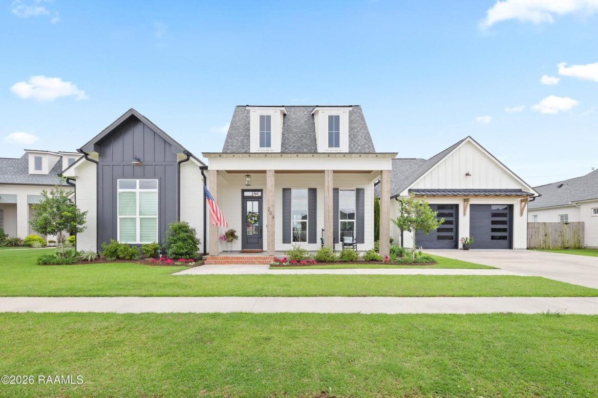 A modern single-story house with a gray and white exterior, a covered front porch with columns, a manicured lawn, an American flag, and a driveway leading to a two-car garage.