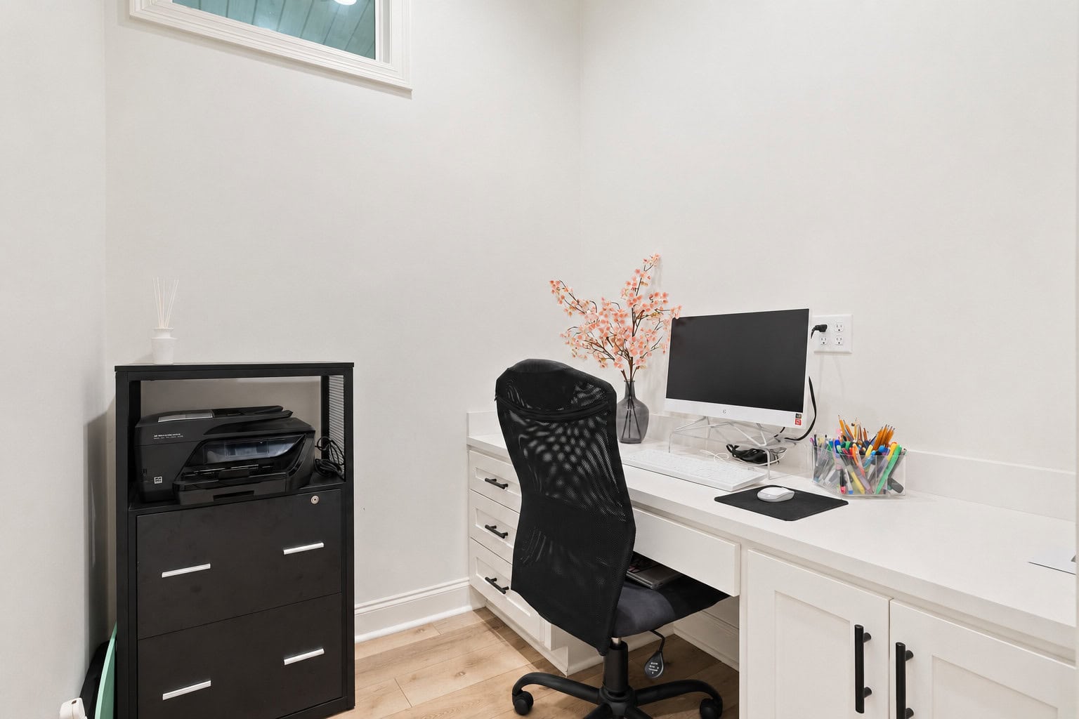 A home office with a white desk, black office chair, desktop computer, printer on a black filing cabinet, a vase with pink flowers, and assorted office supplies on the counter.