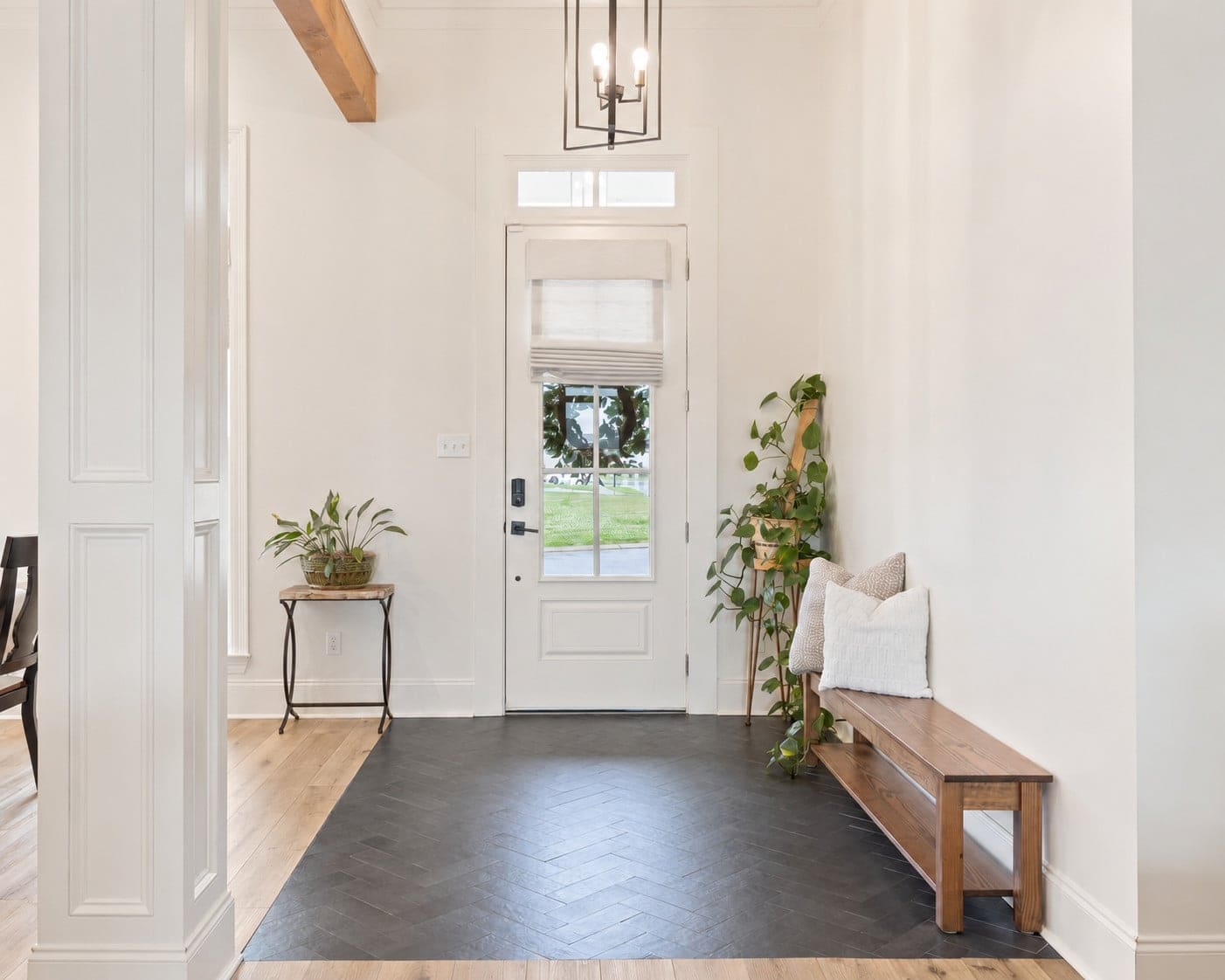 A white entryway with a black herringbone tile floor features a wooden bench with pillows and a plant, a small side table with another plant, and a white door with a window covered by a beige shade.