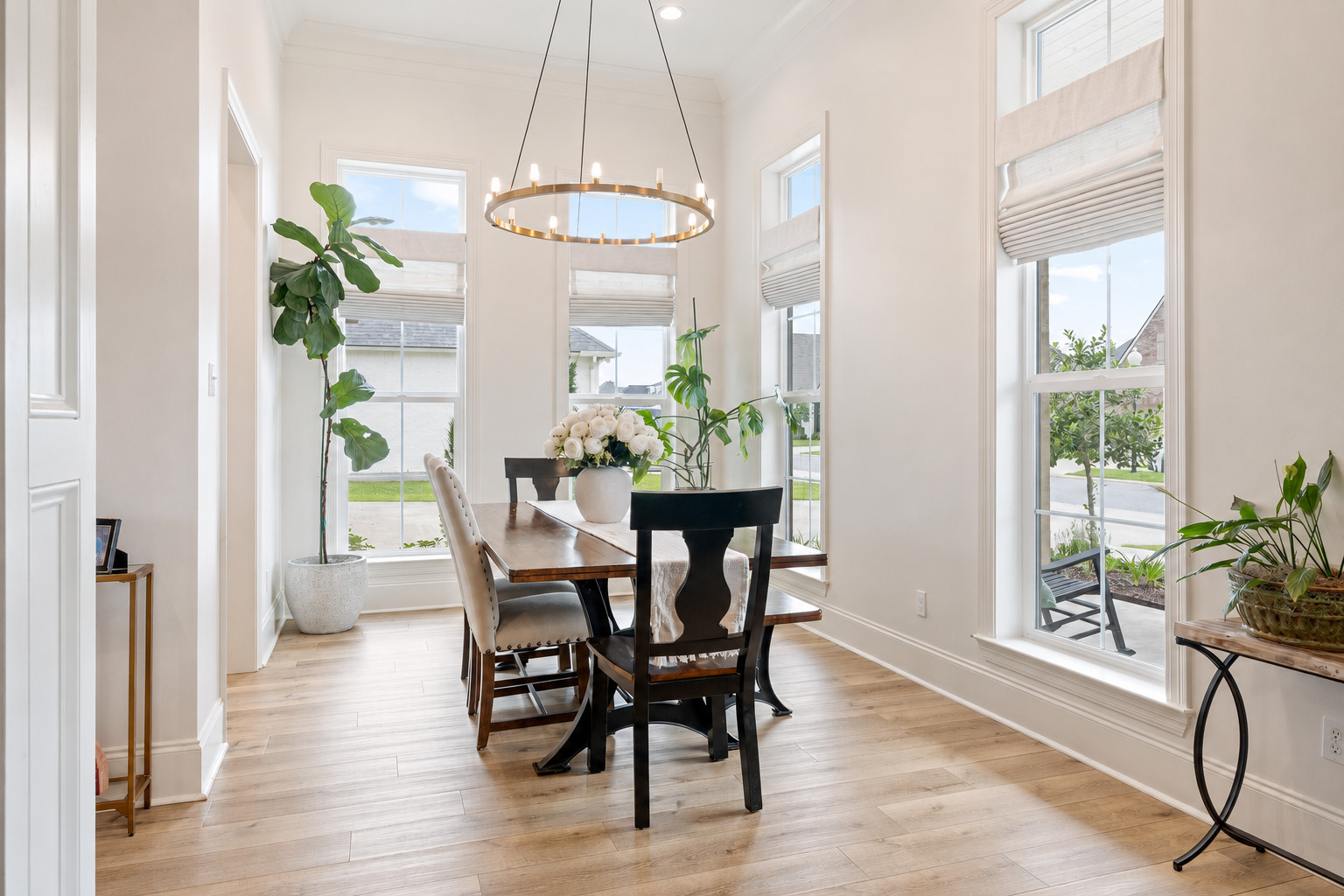 A bright dining room with a wooden table, six chairs, a modern circular chandelier, potted plants, and large windows with white blinds allowing natural light into the space.