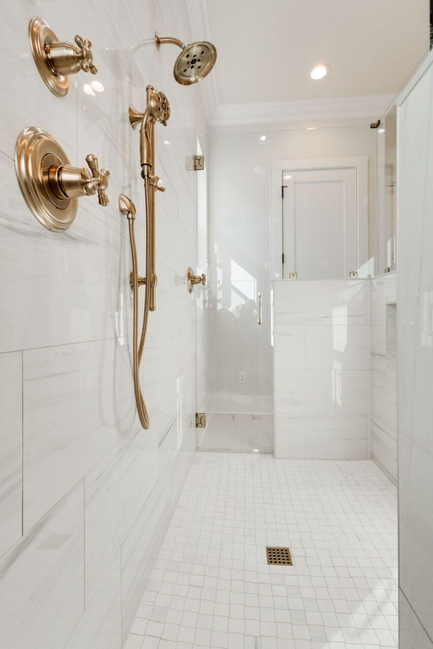 A modern shower with gold fixtures, a handheld showerhead, and a rainfall showerhead. The walls and floor are tiled in white, with a small square drain in the center of the floor.