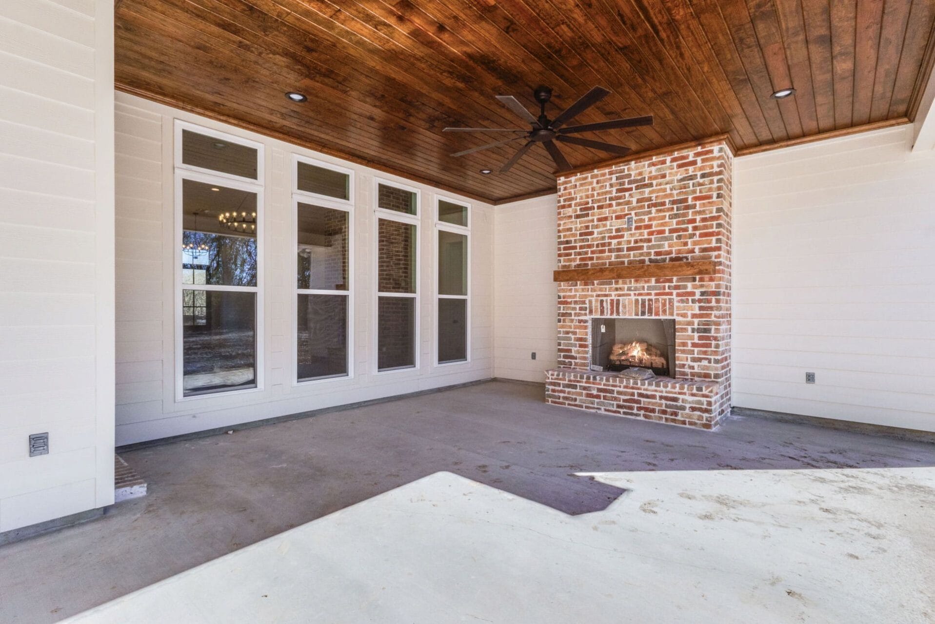 A covered patio with a wooden ceiling, large black ceiling fan, brick fireplace, white paneled walls, and multiple tall windows. The floor is unfinished concrete.