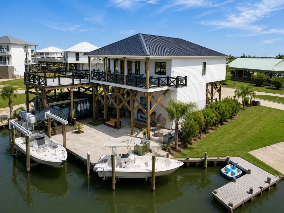 A two-story white house on wooden stilts overlooks a canal with two motorboats and a small pedal boat docked. The house has a deck, balcony, and manicured lawn surrounded by similar homes under a blue sky.