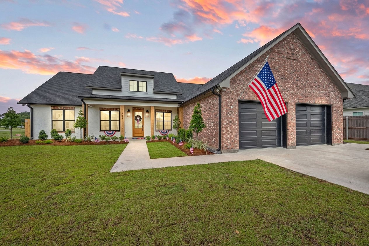 A brick and siding house with two black garage doors, an American flag above the driveway, a wide front lawn, and a sunset sky in the background. Patriotic bunting decorates the porch railing.