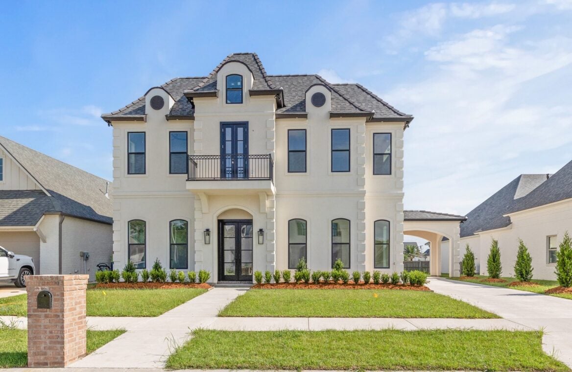A two-story, cream-colored house with black-framed windows, a central balcony, arched front door, and a driveway running under an attached carport. The yard is landscaped with grass, shrubs, and a brick mailbox.