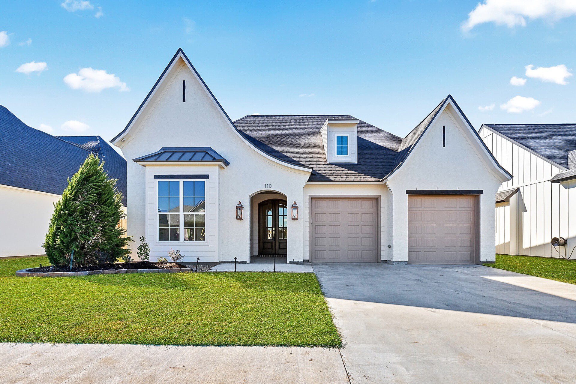 A modern single-story house with a light exterior, two-car garage, arched entryway, landscaped front yard, and a driveway under a clear blue sky.