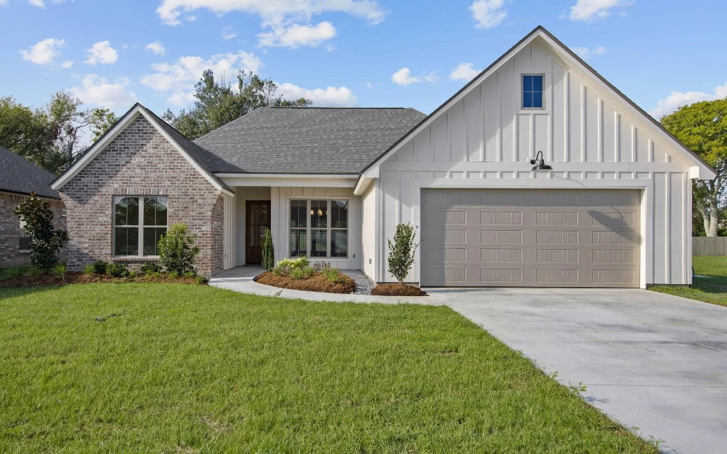 Single-story suburban house with combination brick and vertical siding, a gray shingle roof, double garage door, landscaped front yard, small front porch, and concrete driveway, with blue sky and scattered clouds in the background.