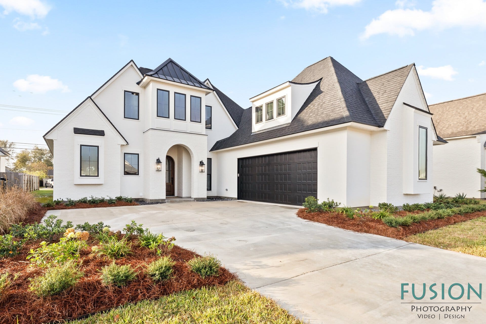 A modern white two-story house with black trim, gabled roofs, large windows, and a double garage. The front yard features a concrete driveway, landscaped garden beds, and manicured shrubs under a clear sky.