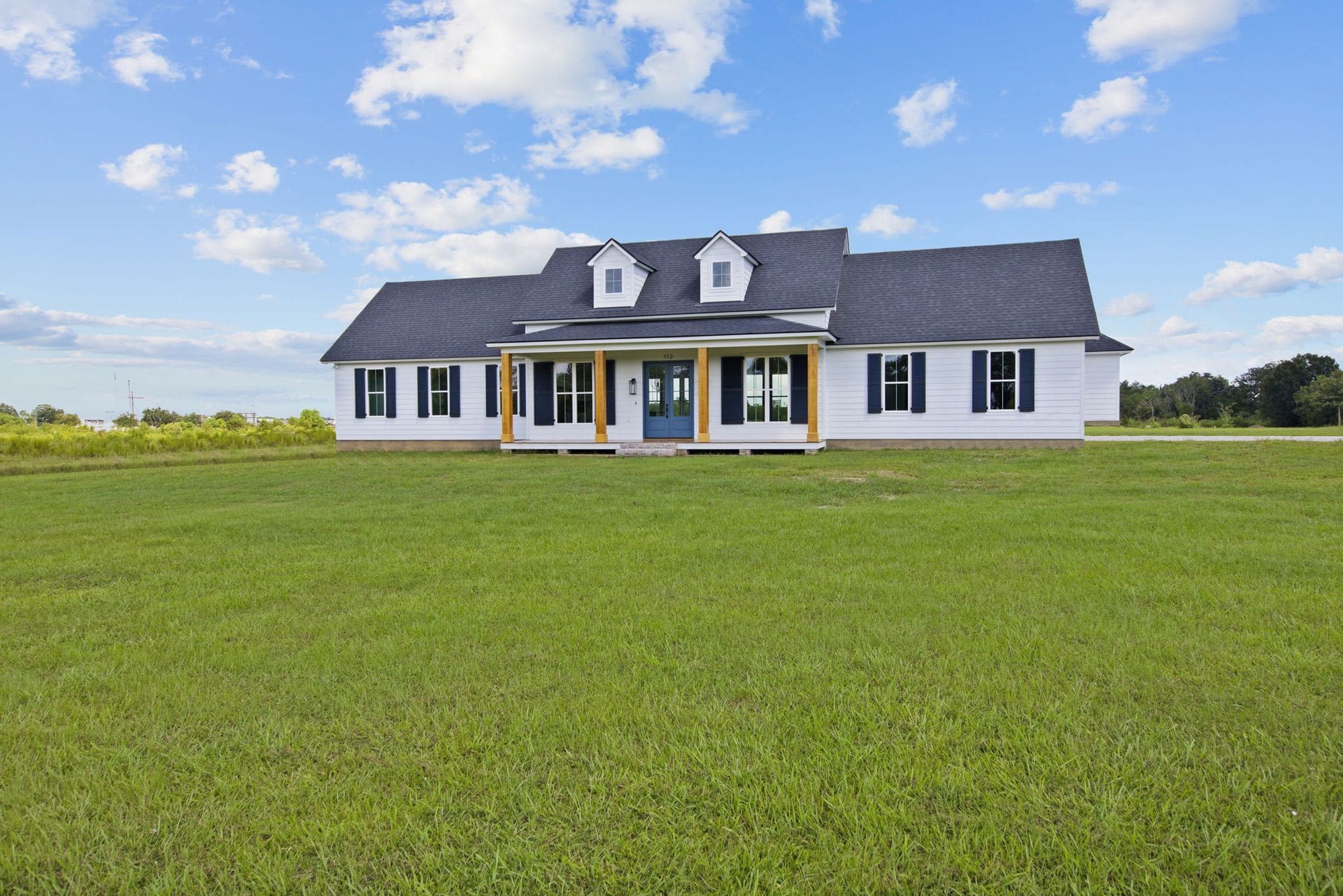 Single-story white house with a dark roof and two small dormer windows, featuring a covered front porch, sits on a large grassy lawn under a partly cloudy sky.