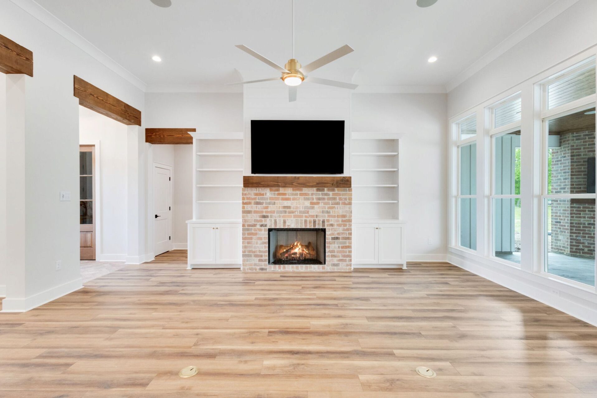 A living room with light wood floors, a brick fireplace with a television mounted above, and built-in white bookshelves.