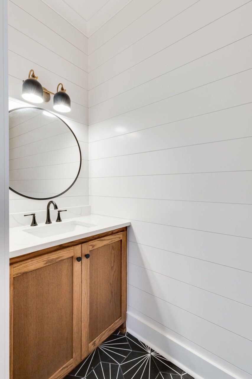 A small bathroom with a wooden vanity, white countertop, round black-framed mirror, and white horizontal wood-paneled walls above a black and white geometric patterned floor.