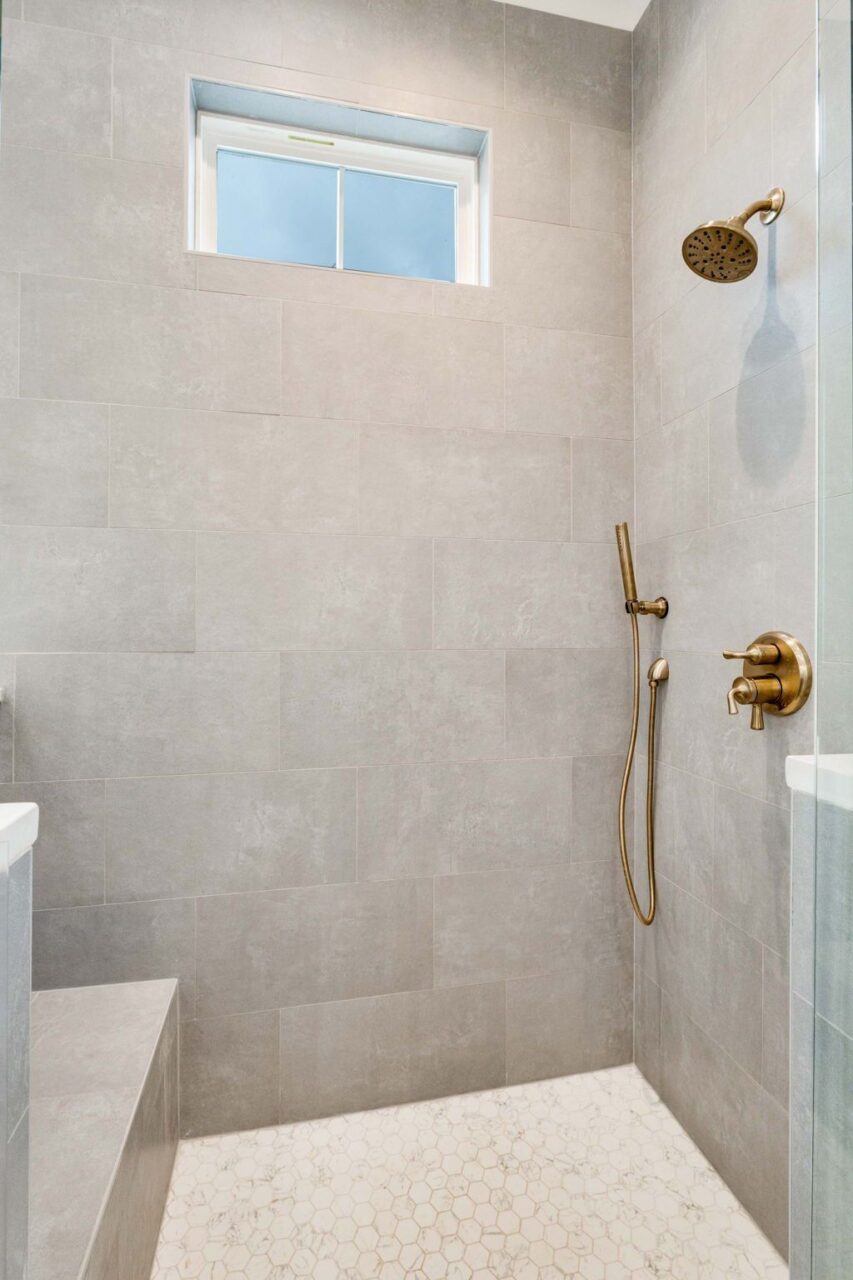 Walk-in shower with grey rectangular wall tiles, a white hexagonal floor, and brass fixtures, including a rainfall showerhead and a handheld sprayer.