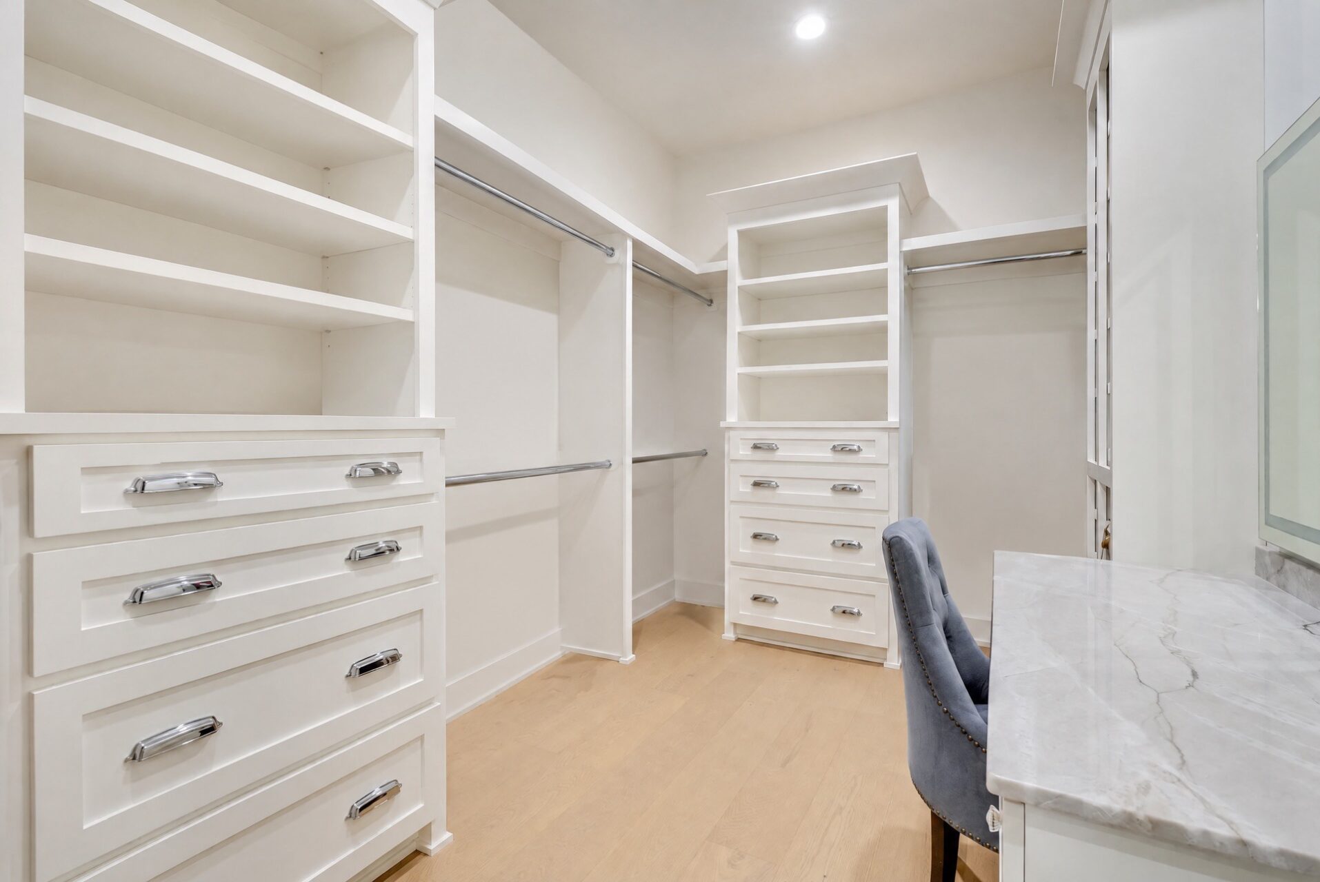 A white walk-in closet with built-in shelves, hanging rods, drawers, and a marble-topped vanity with a tufted gray chair.