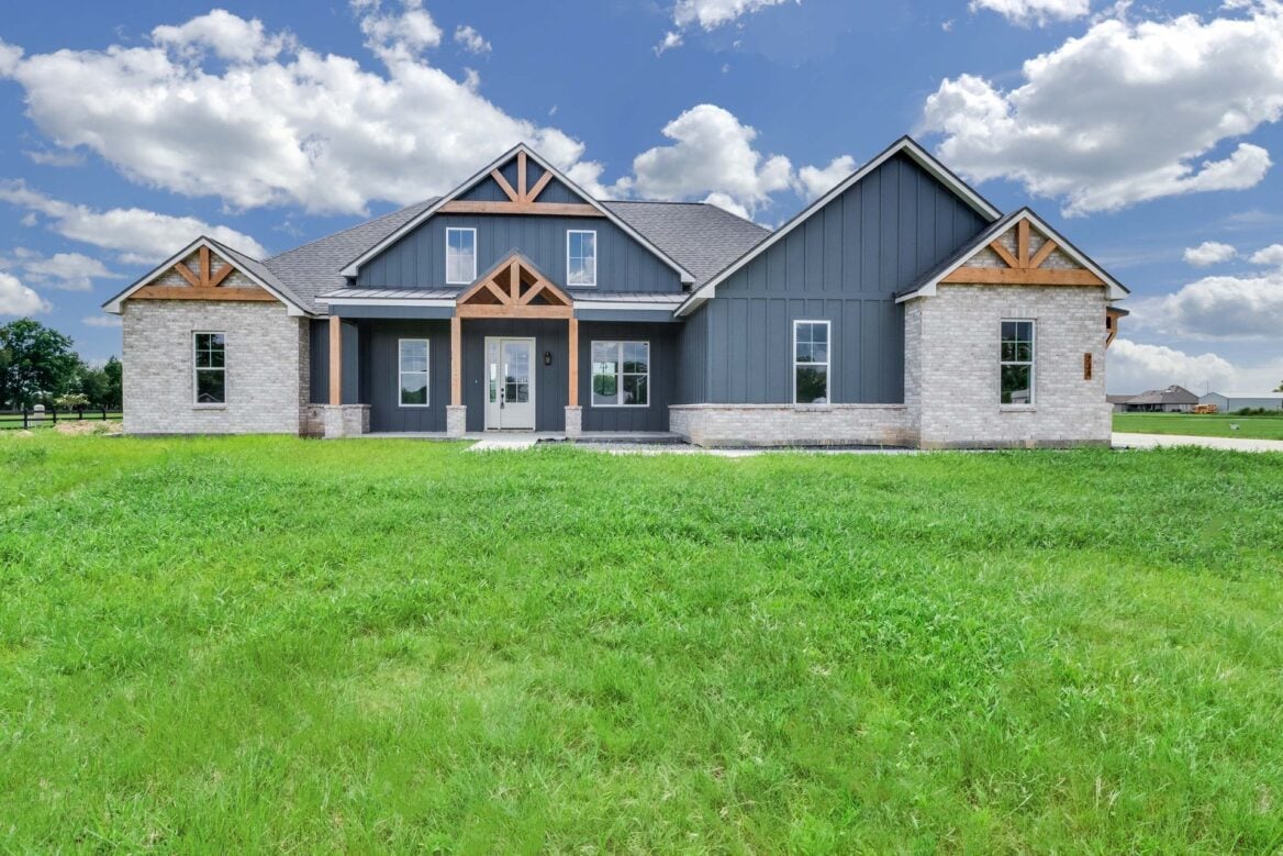 A modern farmhouse with grey vertical siding, white brick accents, and wooden gable brackets, set behind a large grassy yard under a blue sky with clouds.