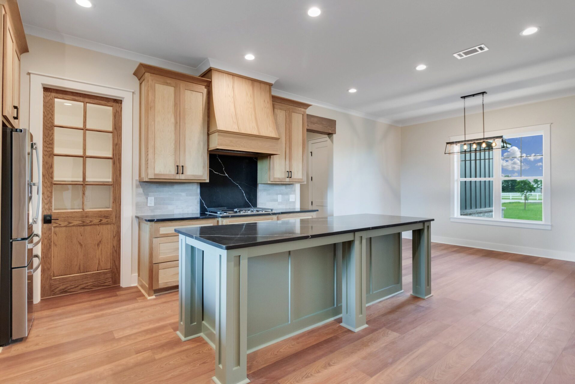 A kitchen with light wood cabinets, a black marble backsplash, and a sage green kitchen island with a black countertop.