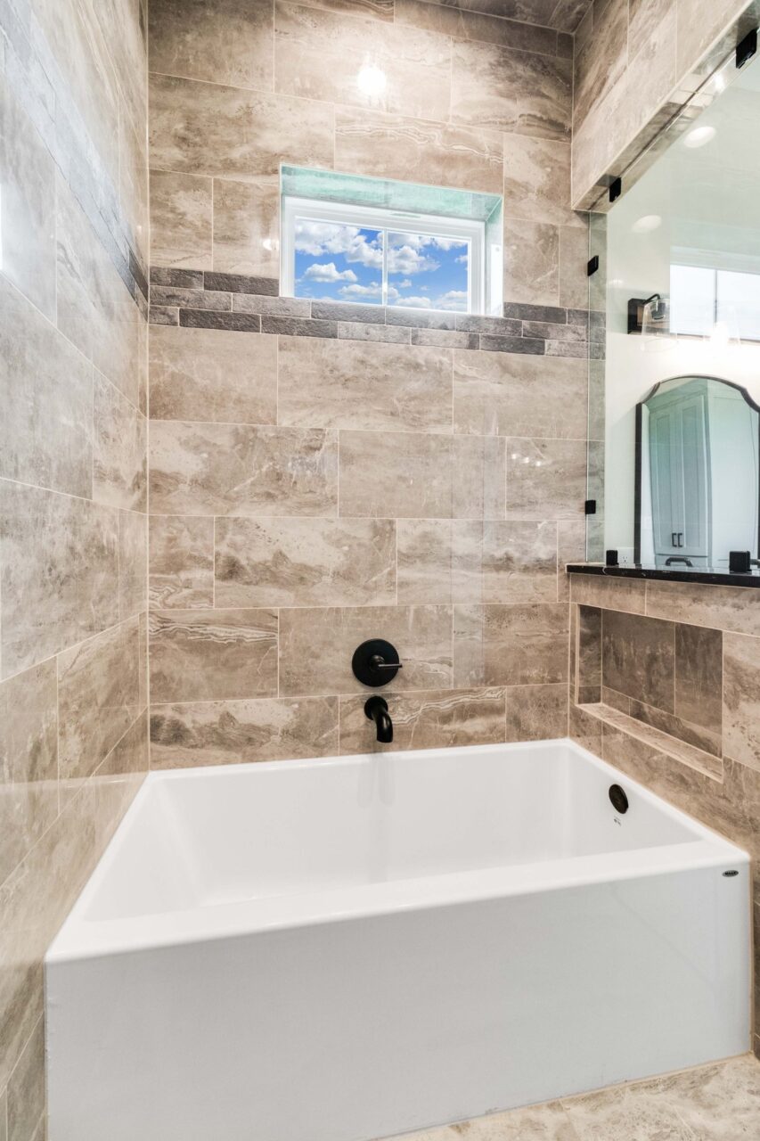 A white bathtub installed against a wall tiled in stone-look porcelain, featuring a small window showing blue sky and black fixtures.