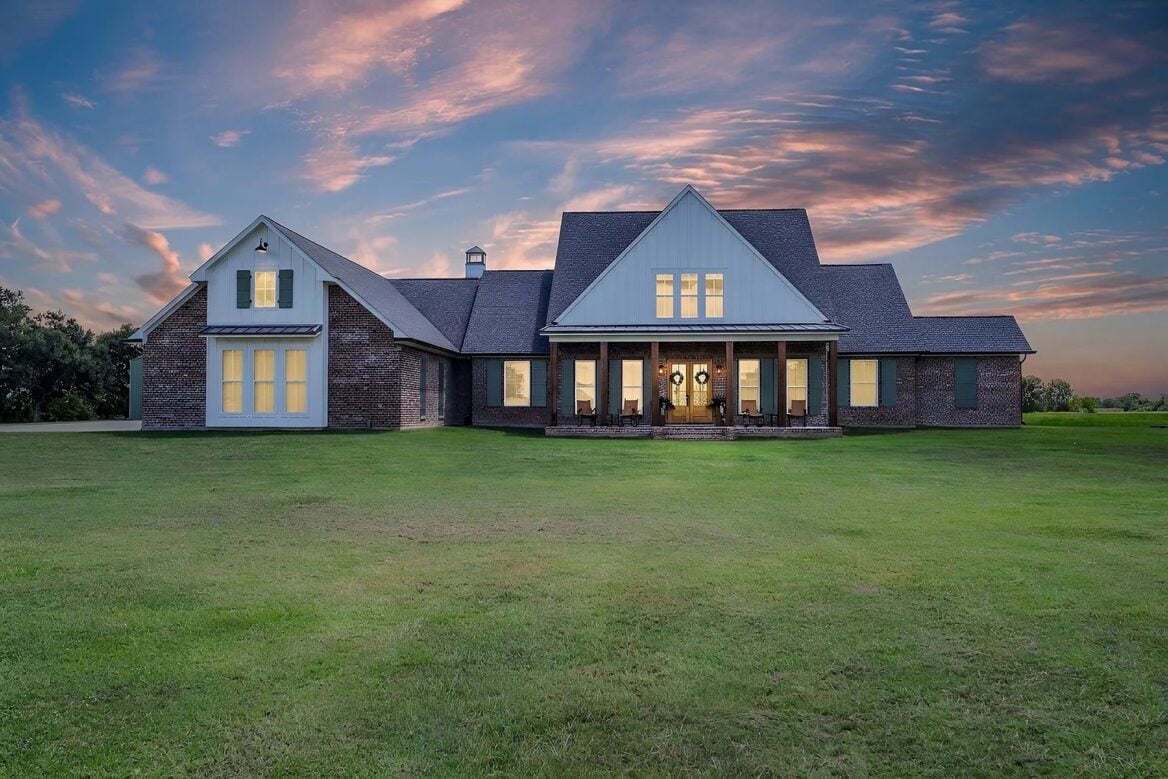 A one-story brick house with white gables and a covered front porch sits in a large grass yard under a sunset sky.