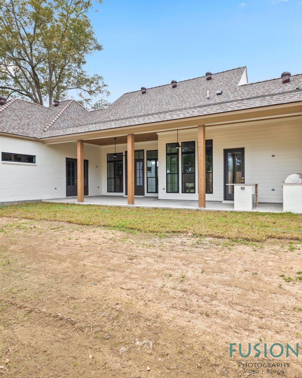 The rear exterior of a white brick house with a covered patio, wooden support columns, and large black-framed windows and doors, overlooking a patchy lawn.