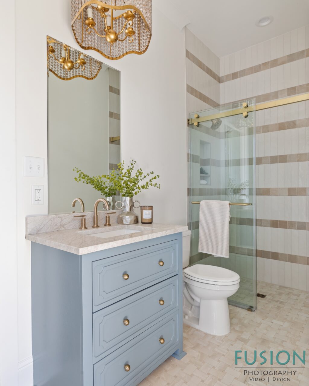 A bathroom featuring a blue vanity with a stone countertop, a gold-trimmed glass shower, and a gold-beaded chandelier.