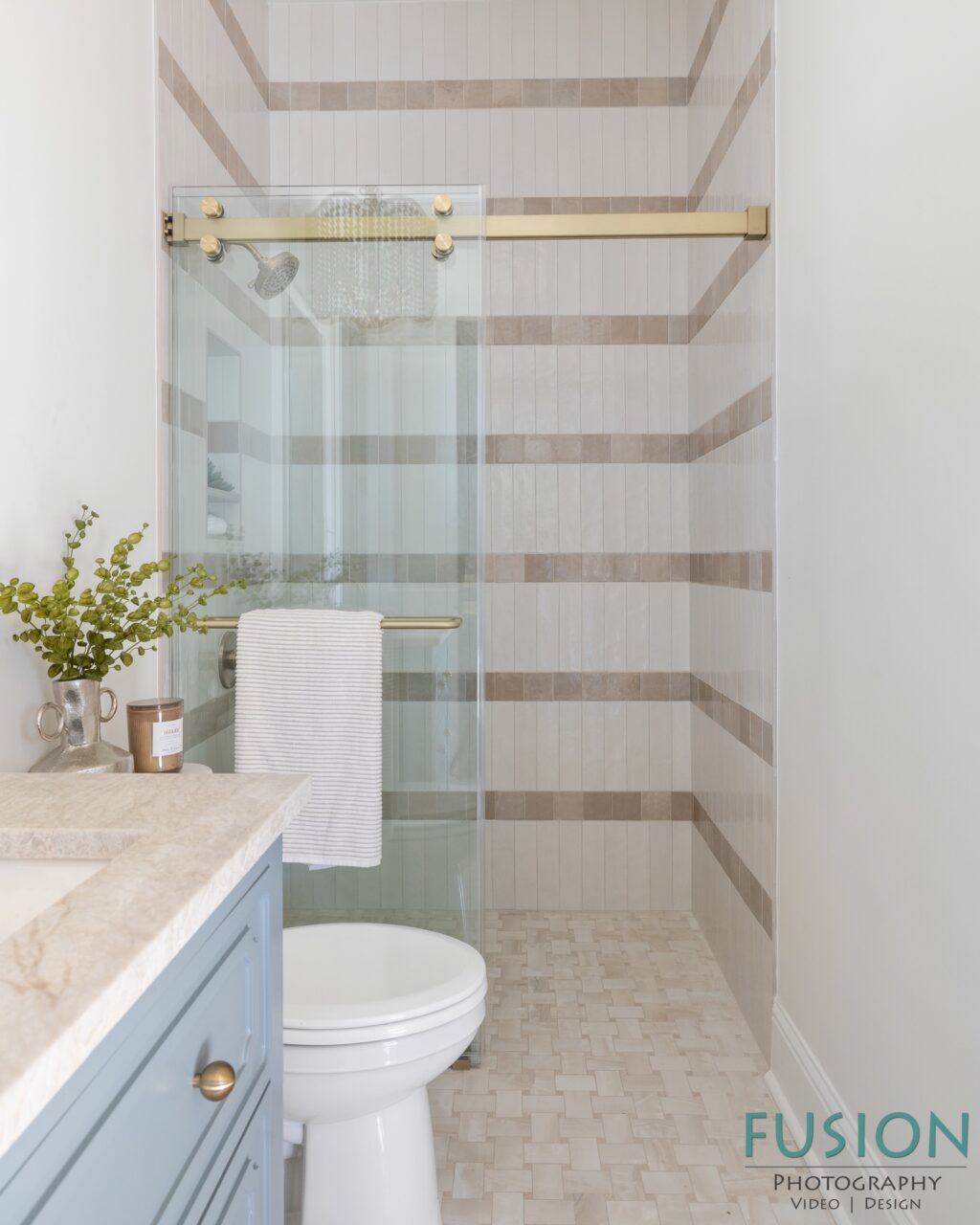 A bathroom with a blue vanity, a toilet, and a glass-enclosed shower featuring horizontal striped tile walls.