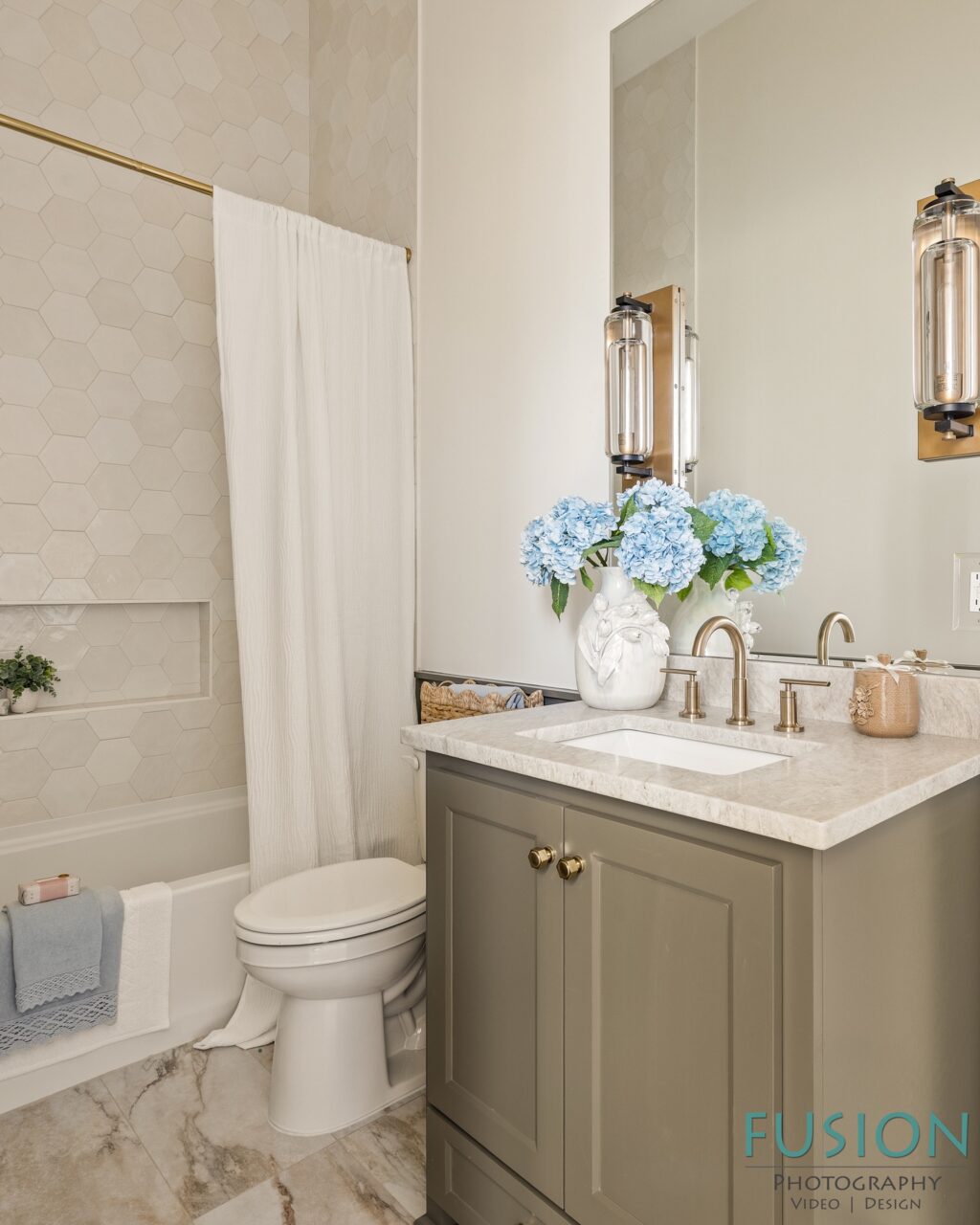 A bathroom featuring a gray vanity with a stone countertop, a white toilet, and a bathtub with a white shower curtain and beige hexagonal wall tiles.