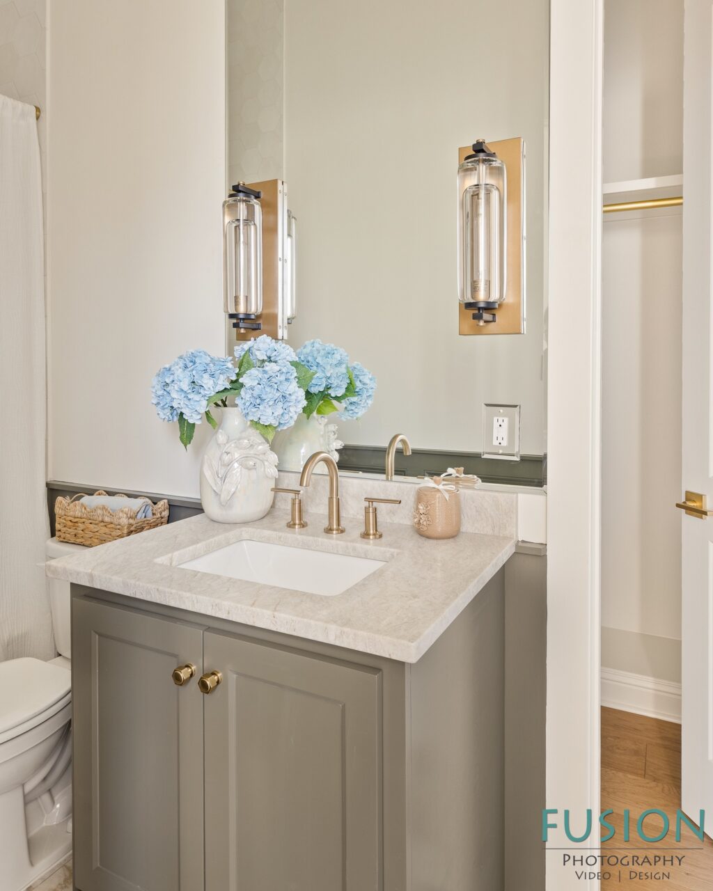 Bathroom vanity with a stone countertop, brass faucet, and a vase of blue hydrangeas in front of a mirror with wall-mounted glass sconces.