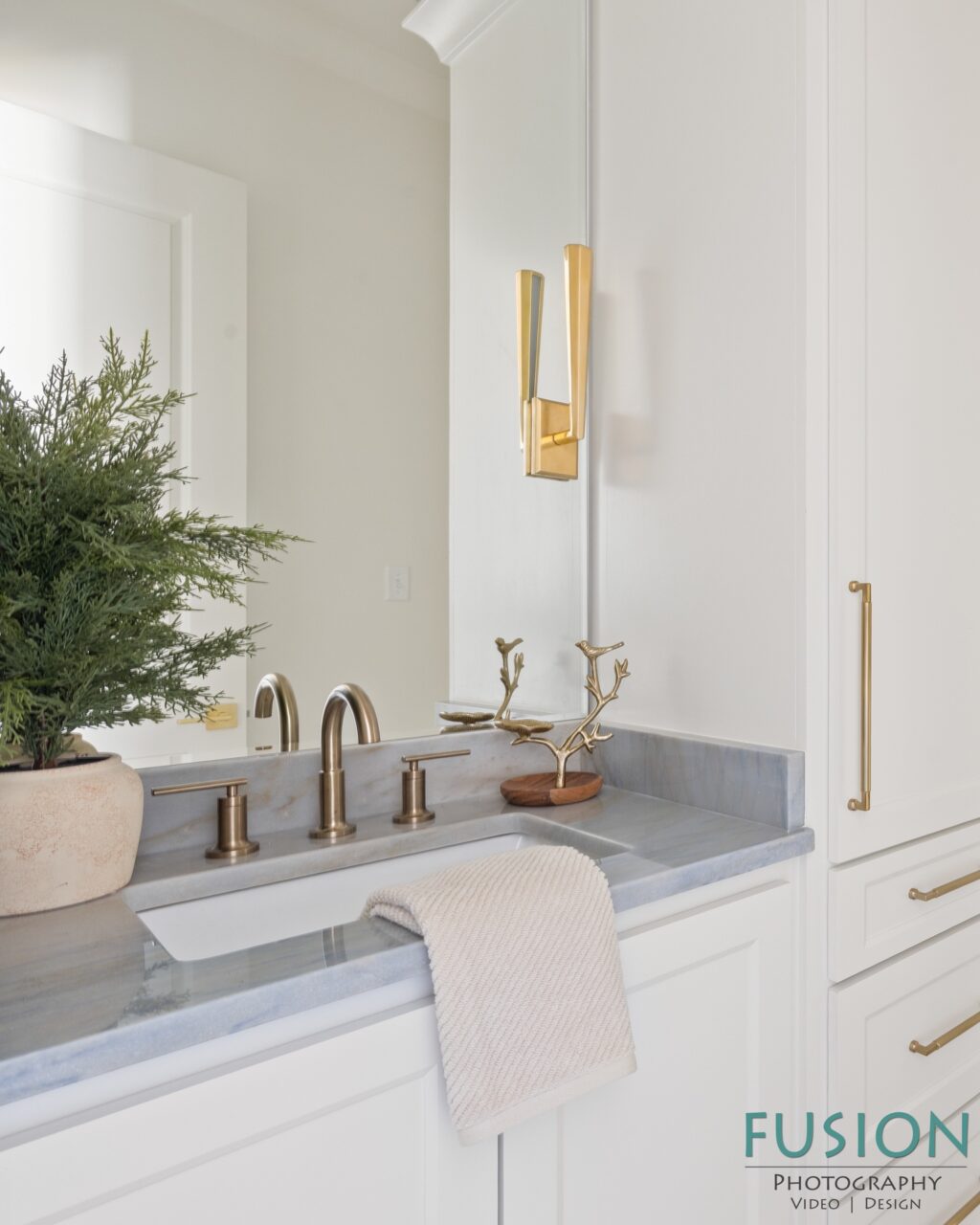 A bathroom vanity with a white countertop, gold faucet, a potted plant, and a decorative bird ornament.
