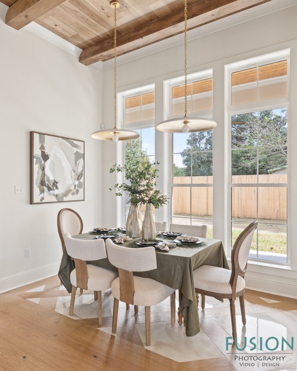 Dining area with a table covered in a green cloth, surrounded by four upholstered chairs, positioned in front of three large windows.