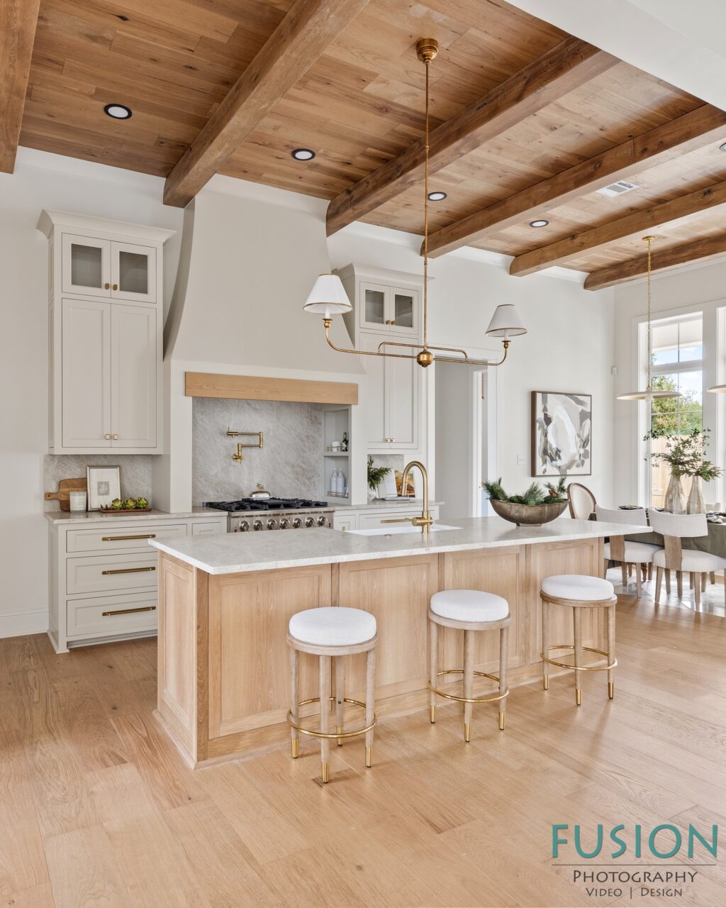 A kitchen featuring a light wood island with three stools, white cabinetry, a marble backsplash, and wooden ceiling beams.