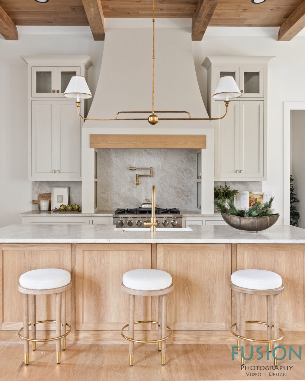 Kitchen island with three white-cushioned stools facing a stove with a range hood and a brass pendant light.