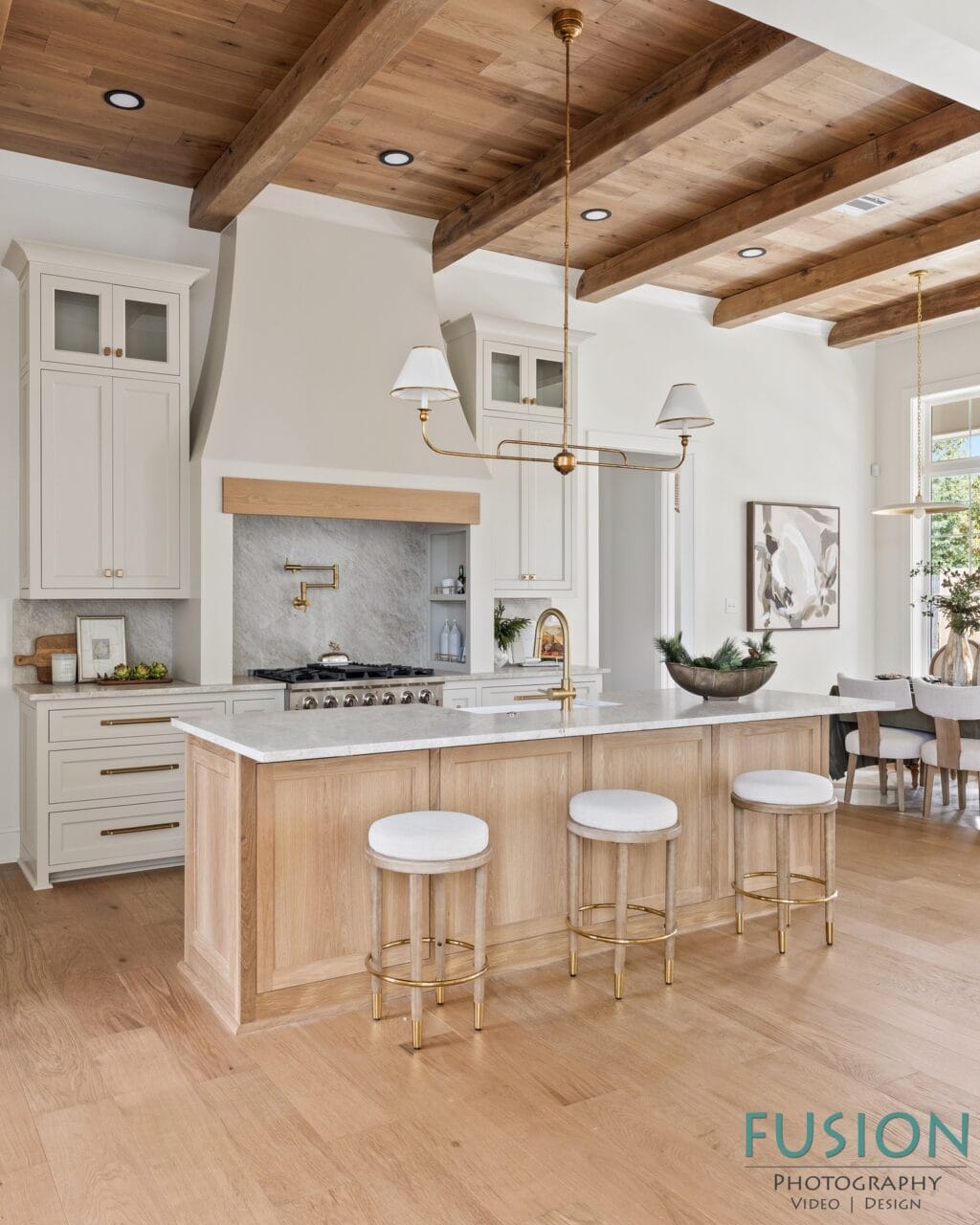 A kitchen with light wood cabinetry, a marble-topped island with three bar stools, and wooden ceiling beams.