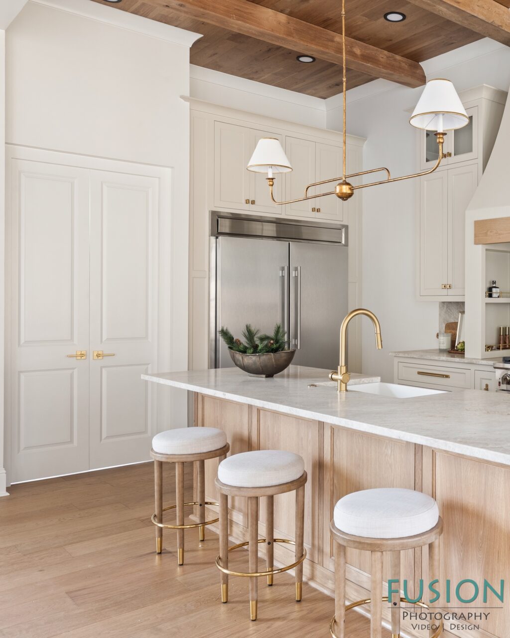 A modern kitchen with a large marble island, three wooden stools with white cushions, a brass faucet, and a brass light fixture suspended from a wood-paneled ceiling.