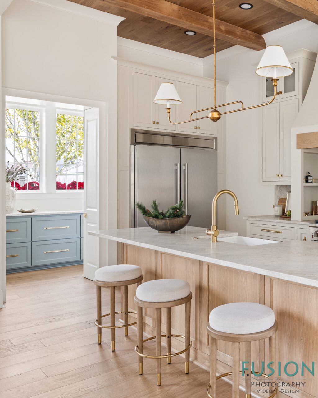 A kitchen island with a stone countertop and three stools in front of a stainless steel refrigerator and light wood cabinets.