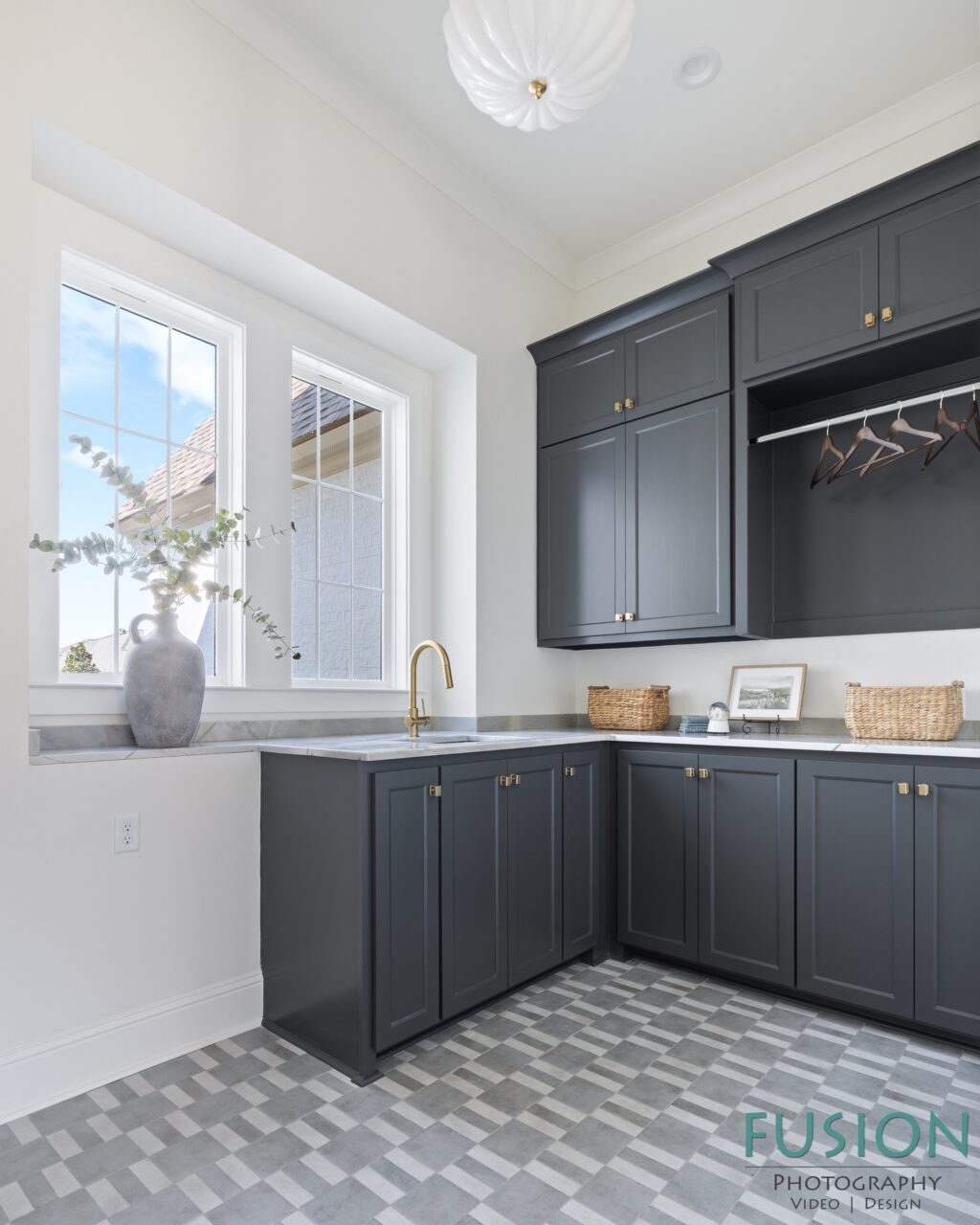 A laundry room features dark grey cabinets, a white countertop with a brass faucet, and a grey and white checkered tiled floor.