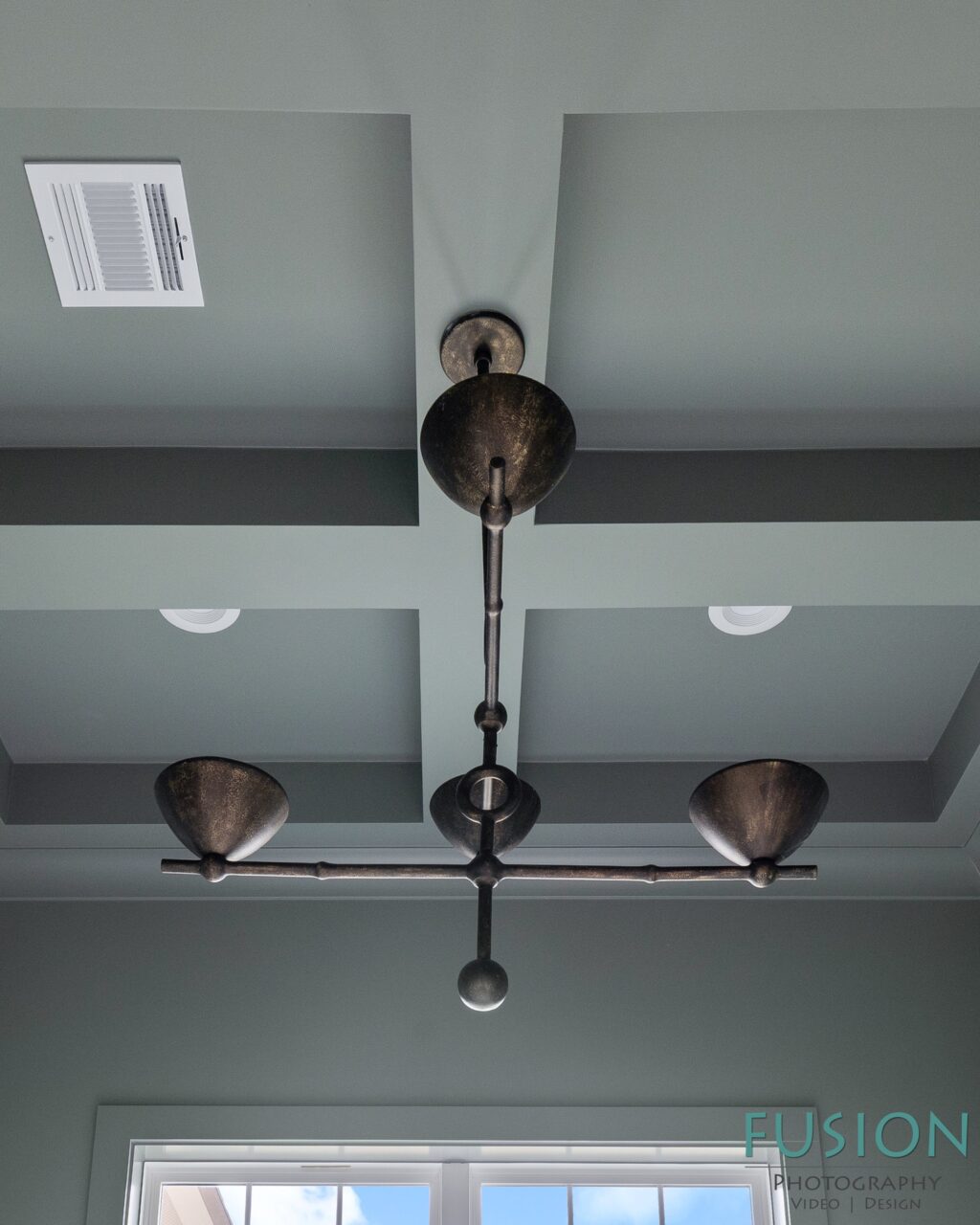 A bronze-finish, multi-arm chandelier hangs from a coffered ceiling painted light blue-gray.