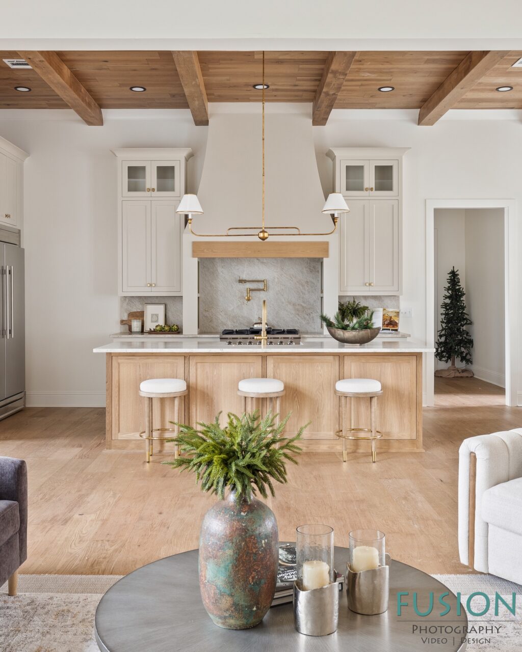 A kitchen with a wood island, white cabinets, a stone backsplash, and a brass range hood above a cooktop, with a circular table and vase in the foreground.