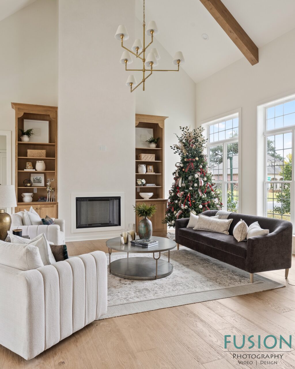 Living room with a fireplace, a decorated Christmas tree, a dark sofa, and two white armchairs arranged around a round coffee table.