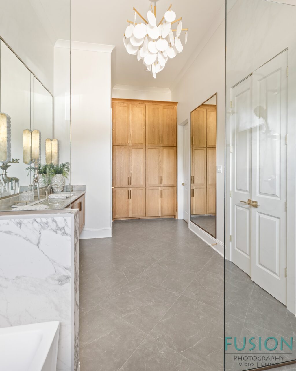 A bathroom featuring a light wood cabinetry unit, a marble-topped vanity with a mirror, and a white chandelier in the center of the room.