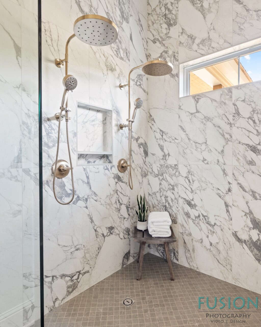 A walk-in shower with white marble walls, gold hardware, dual showerheads, and a small wooden stool holding a stack of folded towels and a plant.