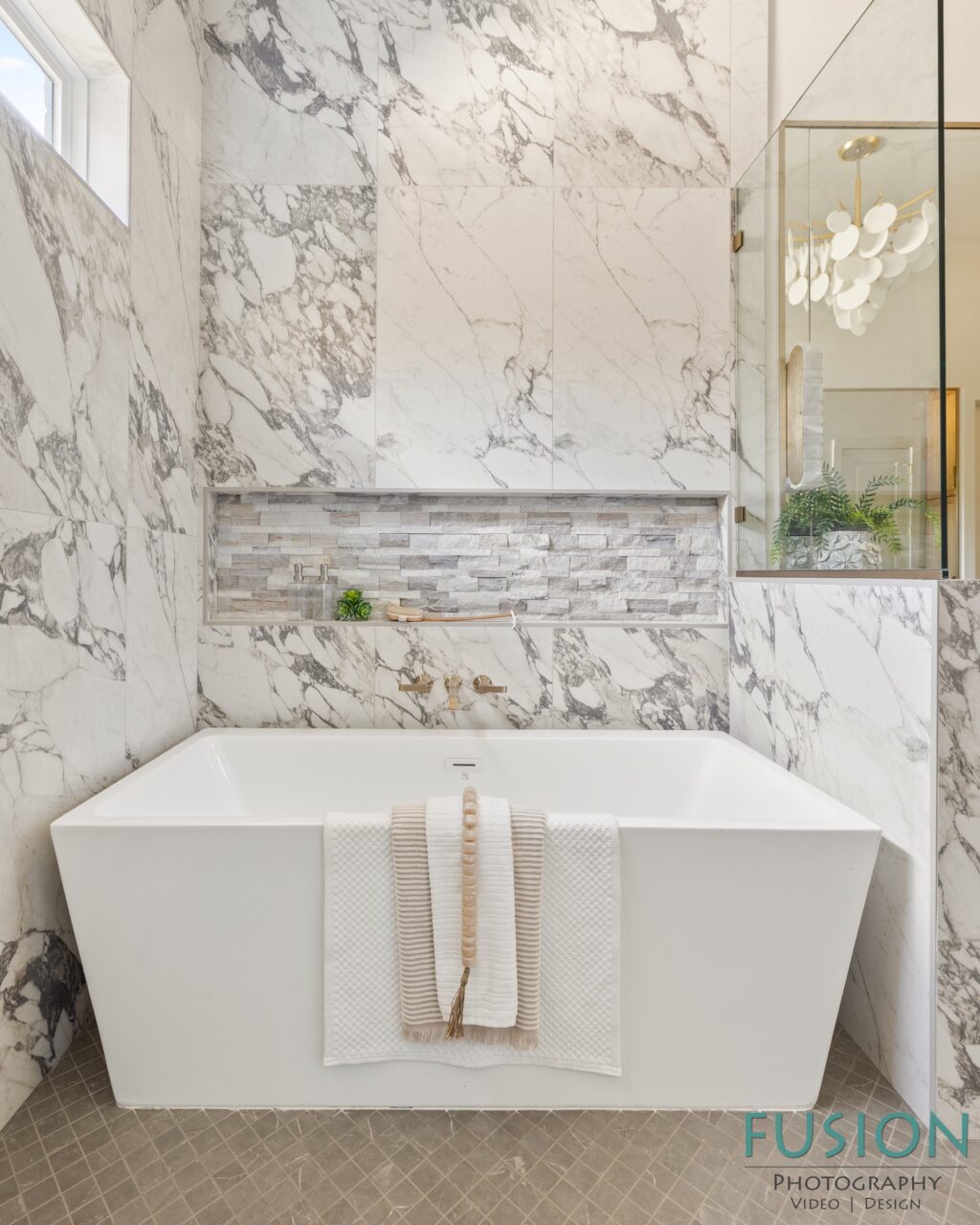 A freestanding white bathtub sits against a marble-tiled wall with a recessed shelf, decorated with a fringed towel and wooden bead strand.