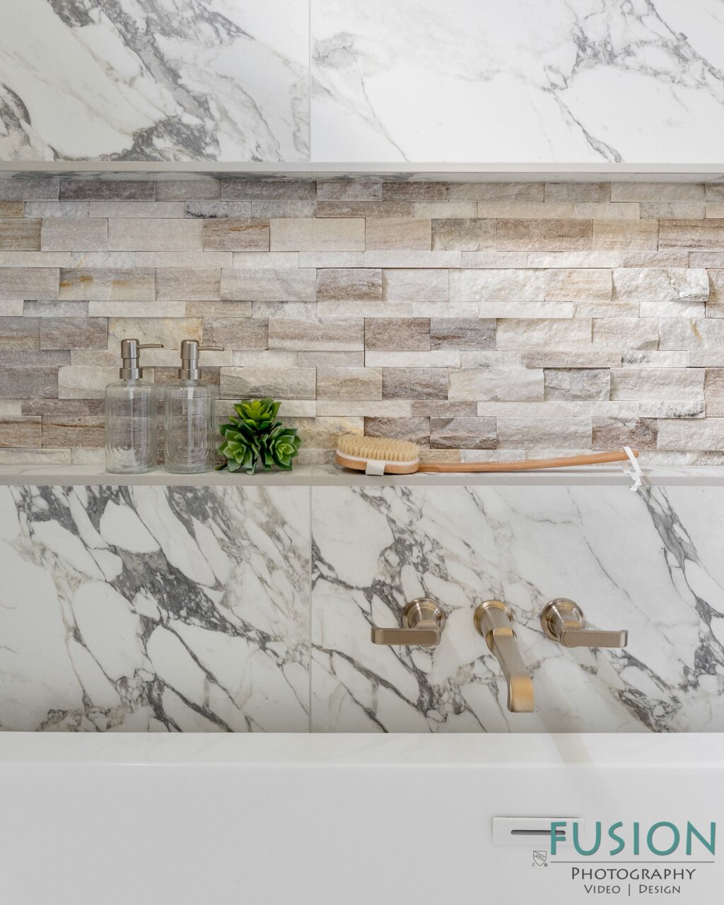 A bathtub ledge features marble tiling, two glass soap dispensers, a small artificial succulent, and a wooden bath brush next to wall-mounted faucet handles.