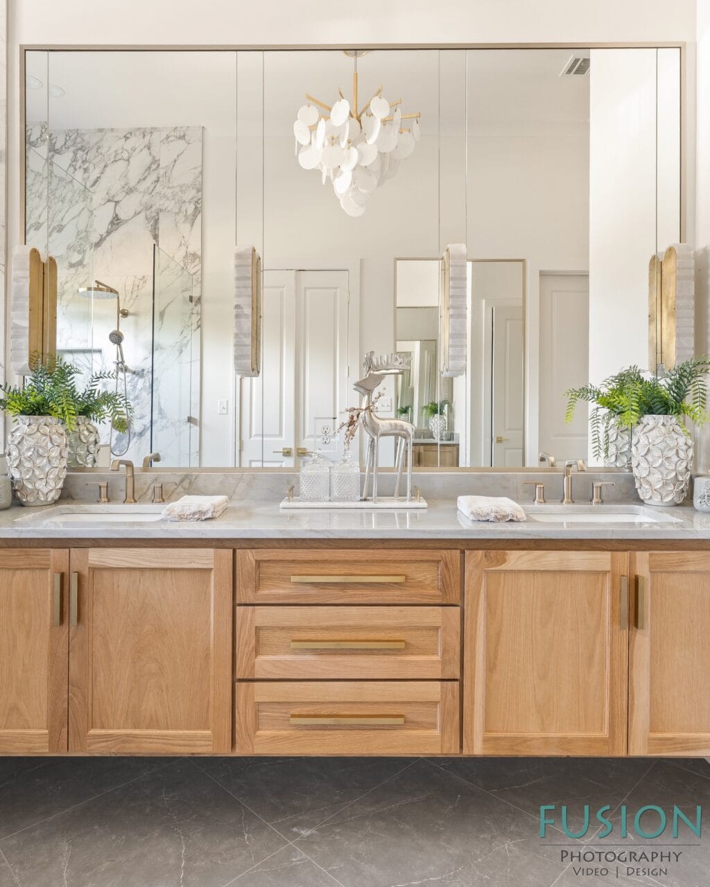 A bathroom vanity with double sinks, light wood cabinetry, brass hardware, and two large potted plants flanking a decorative deer figurine on the countertop.