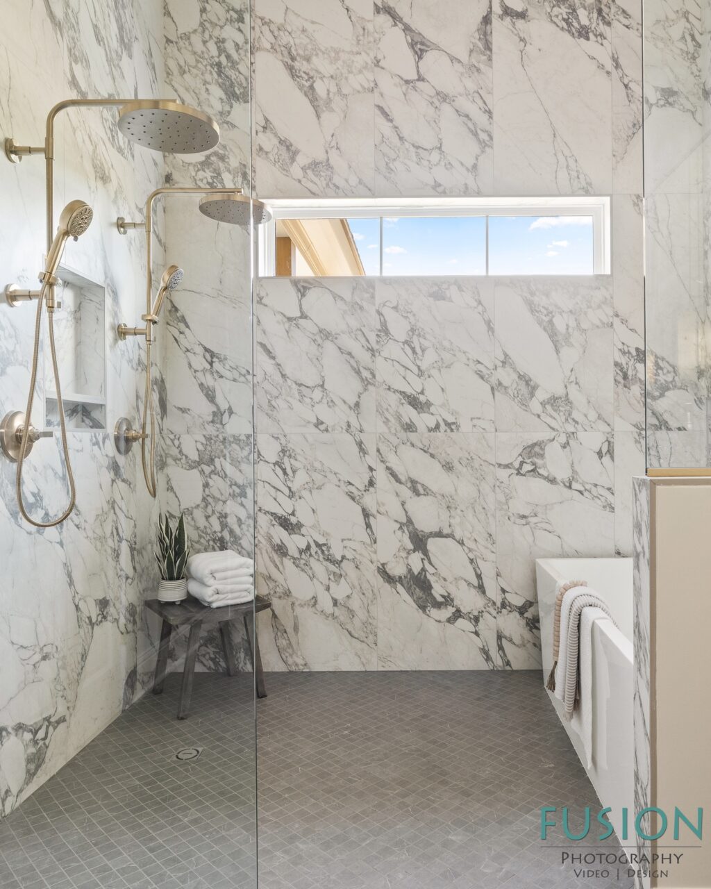 A walk-in shower with marble walls, gold fixtures, a gray tile floor, and a small stool holding folded white towels.