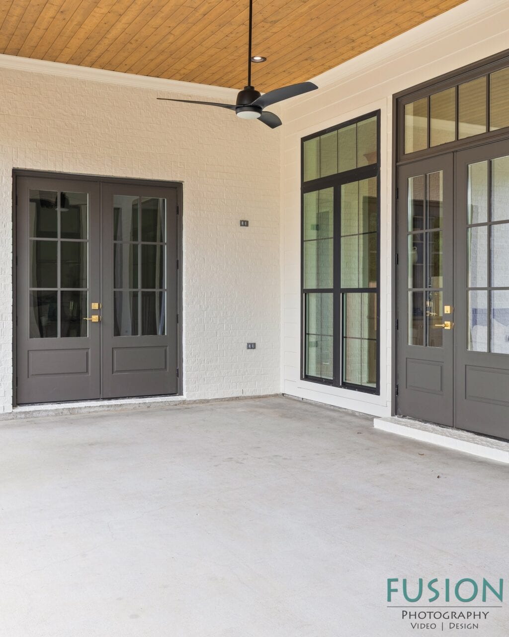 Covered porch with a concrete floor, two sets of dark-framed glass doors, and a ceiling fan against a wood-paneled ceiling.
